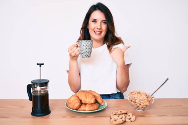Genç, güzel, esmer bir kadın kahvaltı yapıyor. Baş parmağını yana doğru kaldırıyor. Ağzını açıp gülümsüyor. 