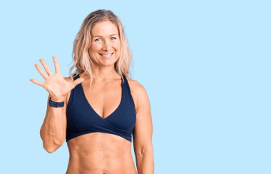 Orta yaşlı, formda, bikini giyen sarışın bir kadın beş numarayı gösteriyor ve gülümsüyor. Kendinden emin ve mutlu.. 