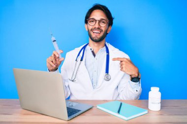 Klinikteki yakışıklı İspanyol doktor elinde şırıngayla gülümseyen mutlu ve gururlu birini işaret ediyor. 