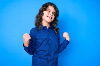 Uzun saçlı sevimli İspanyol çocuk gündelik giysiler giyiyor. Çok mutlu ve heyecanlı. Kollarını kaldırıp, gülümseyerek ve başarı için bağırarak kazanan jestler yapıyor. kutlama konsepti. 