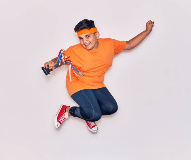 Spor kıyafetleri giyen sevimli İspanyol çocuk mutlu gülümsüyor. Yüzünde tebessümle zıplıyor İzole edilmiş beyaz arka planda ödül tutuyordu