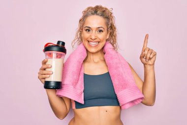 Güzel sarışın sporcu kadın havluyla protein içeceği tutuyor. Bir fikir ya da soru işareti ile şaşırmış, mutlu yüzlü, bir numara.