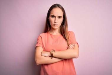 İzole edilmiş pembe arka plan üzerinde duran sıradan tişört giyen genç ve güzel bir kadın şüpheci ve gergin, yüzünde çapraz kollu tasvip etmeyen bir ifade. Negatif kişi.