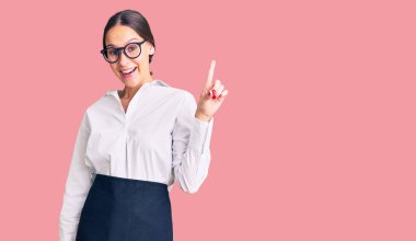 Profesyonel garson önlüğü giymiş güzel esmer genç bir kadın başarılı bir fikirle işaret ediyor. Heyecanlı ve mutlu. Bir numara.. 