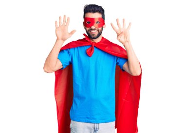 Sakallı, süper kahraman kostümü giyen genç ve yakışıklı bir adam dokuz numaralı parmaklarını gösterip gülümserken kendinden emin ve mutlu.. 