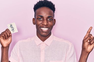 Elinde dolar sembolü olan genç Afrikalı Amerikalı adam gülümseyen mutlu bir şekilde elini ve parmağını kenarı işaret ediyor. 