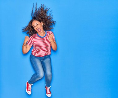 Orta yaşlı, güzel, İspanyol bir kadın. Günlük kıyafetler giyip mutlu gülümsüyor. Yüzünde gülücükle zıplayarak kazanan jestler yaparak izole edilmiş mavi arka planda yumruklar sallayarak