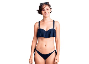 Kısa saçlı güzel bir kadın bikini giyiyor. Pozitif ve mutlu görünüyor. Duruyor ve gülümsüyor. 