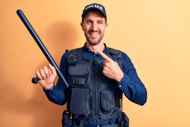 Üniforma giyen yakışıklı bir polis ve sarı arka planda değnek tutan bir kurşunla mutlu bir şekilde elini ve parmağını göstererek gülümsüyor.