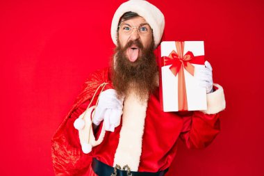 Uzun sakallı, yakışıklı kızıl saçlı Noel Baba kostümü giyen ve hediye dolu büyük çantalı komik bir ifadeyle dilini dışarı çıkaran adam.. 