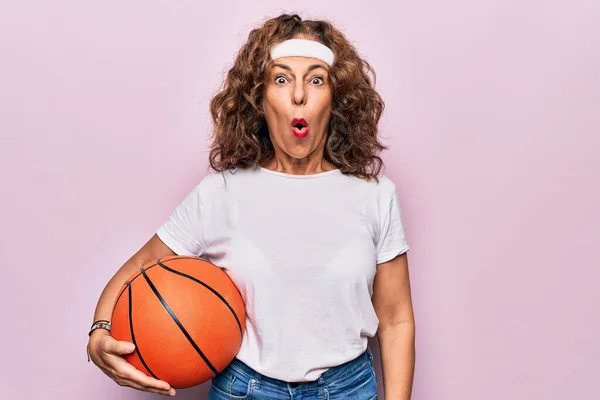 中年黑发女子篮球运动员拿着球在孤立的粉色背景下害怕和惊讶张开嘴惊喜 难以置信的脸 — 图库照片