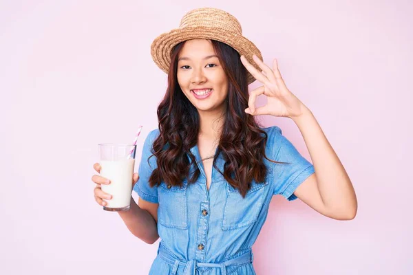 年轻美丽的中国姑娘头戴夏帽 手里拿着一杯牛奶 手拿着手签 微笑着表示友善的手势 象征着极好的象征 — 图库照片