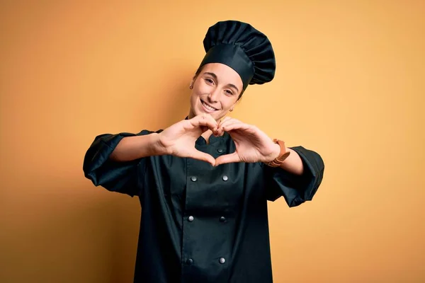 Young beautiful chef woman wearing cooker uniform and hat standing over yellow background smiling in love doing heart symbol shape with hands. Romantic concept.