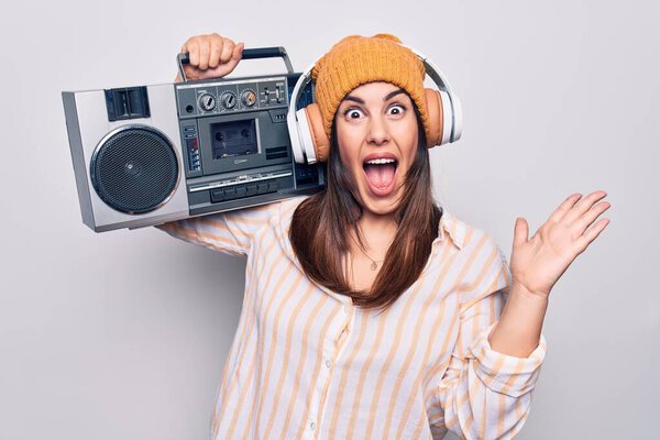 Young beautiful brunette woman listening to music using vintage boombox and headphones celebrating achievement with happy smile and winner expression with raised hand