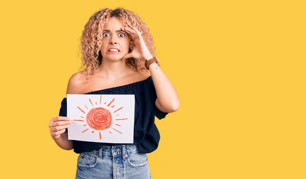 Young blonde woman with curly hair holding sun draw stressed and frustrated with hand on head, surprised and angry face