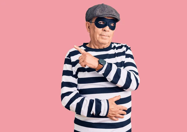 Senior handsome man wearing burglar mask and t-shirt pointing with hand finger to the side showing advertisement, serious and calm face