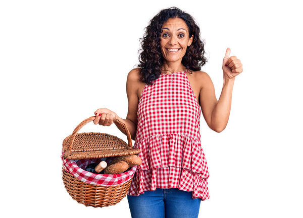 Middle age beautiful woman holding picnic basket with food smiling happy and positive, thumb up doing excellent and approval sign 