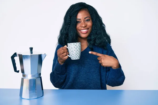 Beautiful african woman drinking a coffee from italian coffee maker smiling happy pointing with hand and finger