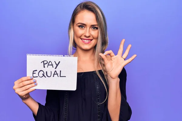 Beautiful blonde woman asking for equality economy holding paper with pay equal message doing ok sign with fingers, smiling friendly gesturing excellent symbol