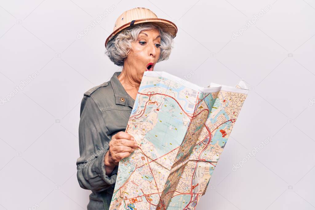 Senior grey-haired woman wearing explorer hat holding map scared and amazed with open mouth for surprise, disbelief face 