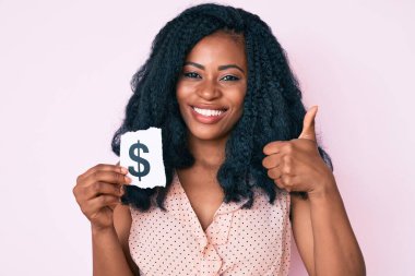 Güzel Afrikalı kadın elinde dolar sembolü kağıdıyla mutlu ve pozitif gülümsüyor, başparmağı mükemmel ve onay işareti yapıyor. 