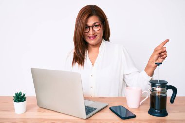 Orta yaşlı esmer, ispanyol iş kadını ofiste çalışıyor. Gülümsüyor, mutlu bir şekilde elini ve parmağını yana doğru gösteriyor. 