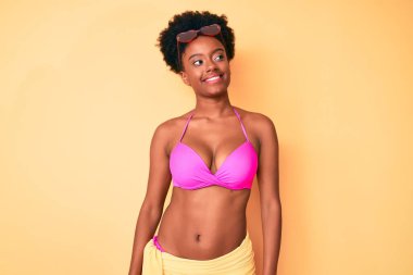 Genç Afro-Amerikan kadın bikini giyiyor. Yüzünde gülümsemeyle yan yana bakıyor. Doğal bir ifade. Kendinden emin gülen. 