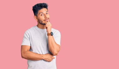 Yakışıklı, gündelik tişört giyen, kameraya kendinden emin, kolları çapraz ve çenesi kalkık bir şekilde bakan Latin Amerikalı bir genç. Olumlu düşünmek. 