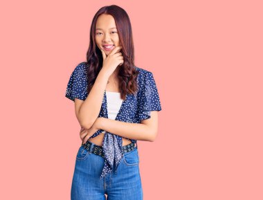 Gündelik kıyafetler giyen genç, güzel Çinli kız kameraya bakıyor kollarını kavuşturup gülümsüyor ve çenesini kaldırıyor. Olumlu düşünmek. 