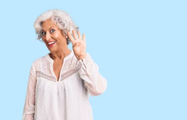 Kıdemli, gri saçlı, günlük kıyafetler giyen, dört numarayı gösteren ve gülümseyen, kendinden emin ve mutlu bir kadın.. 