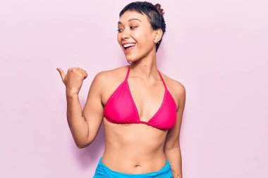 Bikini giyen genç bir kadın başparmağını yana uzatıyor ve ağzı açık gülümsüyor. 