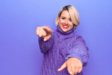 Güzel sarışın ve cüsseli bir kadın mor arka plan üzerine balıkçı yaka kazak giyip seni ve kamerayı parmaklarıyla göstererek gülümsüyor.