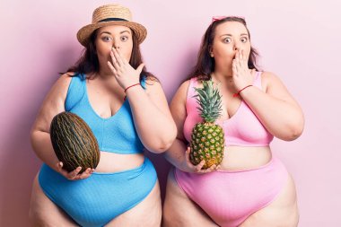 Bikini giyen, kavun tutan ve ağzı ağızla kaplanmış ananas giyen genç ve büyük beden ikizler, hata yapmaktan korkuyorlar. şaşırmış ifade 