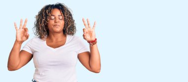 Genç Afro-Amerikan artı cüsseli kadın rahat giyiniyor ve gözleri kapalı gülümsüyor parmaklarıyla meditasyon hareketi yapıyor. yoga konsepti. 