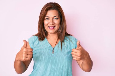 Orta yaşlı esmer, günlük kıyafetler giyen İspanyol kadın. Başarı işareti ile el hareketi yapıyor. Gülümsüyor ve mutlu. Neşeli ifade ve kazanan jest. 