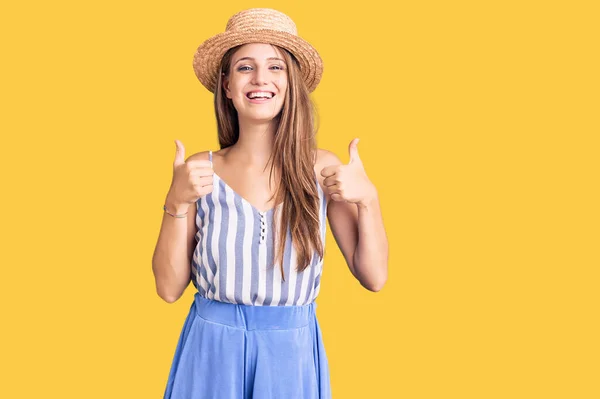 Young beautiful blonde woman wearing summer hat success sign doing positive gesture with hand, thumbs up smiling and happy. cheerful expression and winner gesture.