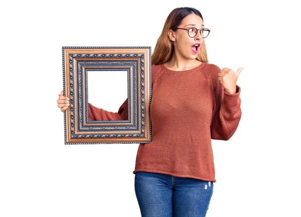 Beautiful Young Woman Holding Empty Frame Pointing Thumb Side Smiling — Stock Photo, Image