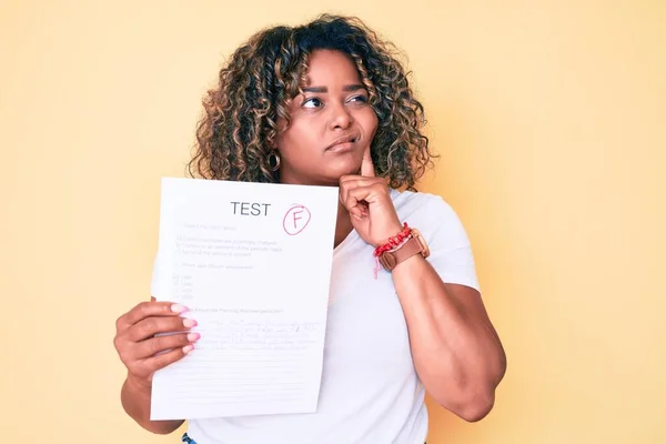 Young african american plus size woman showing a failed exam serious face thinking about question with hand on chin, thoughtful about confusing idea