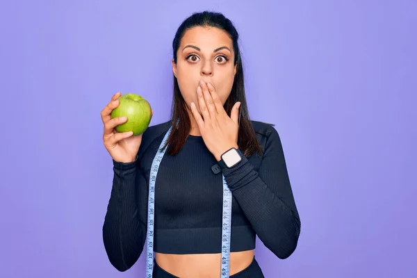 テープを使用して美しいブルネットのスポーティな女性は 手で口をカバー紫色の背景にリンゴの果物を食べる測定ショックを受け 間違いを恐れている 驚きの表情 — ストック写真