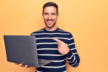 Genç yakışıklı adam dizüstü bilgisayar kullanarak çalışıyor. Sarı arka planda duruyor. Mutlu bir gülümsemeyle işaret ediyor.