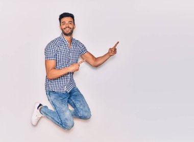 Genç, yakışıklı, sıradan kıyafetler giyen, mutlu gülümseyen bir İspanyol. Yüzünde tebessümle sıçrayıp, izole beyaz arka planda parmaklarını yana doğru gösteriyor.