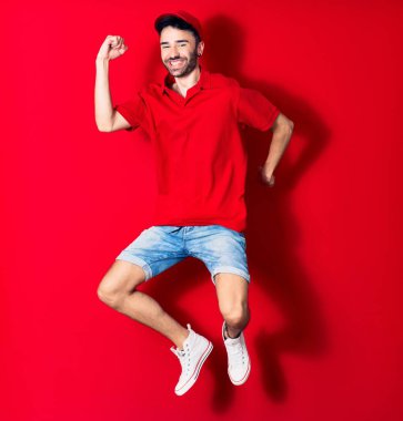 Şapkalı genç teslimatçı mutlu gülümsüyor. İzole edilmiş kırmızı arka planda yüzünde gülümsemeyle zıplıyor