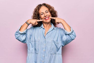 Orta yaşlı güzel bir kadın pembe arka planda dikilmiş sıradan bir kot gömlek giyiyor. Gülümsüyor ve parmaklarıyla ve ağzıyla işaret ediyor. Diş sağlığı konsepti.
