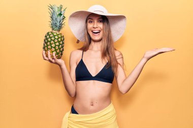 Bikini giyen, şapkalı, ananaslı genç bir kız mutluluğu kutluyor. Gülümsüyor ve elini kaldırarak kazanan yüz ifadesi sergiliyor. 