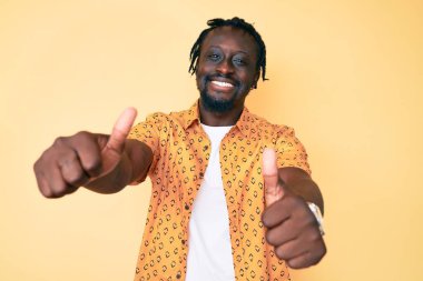 Saçları örgülü, el hareketleriyle olumlu jest yapmayı onaylayan, gülümseyen ve başarı için sevinen Afro-Amerikan genç bir adam. kazanan jest. 
