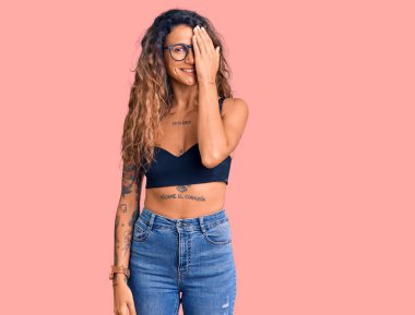 Dövmeli, sıradan kıyafetler giyen, gözlüklü, bir gözünü eliyle kapatan, yüzünde kendine güvenen bir gülümseme ve sürpriz duygular olan İspanyol bir kadın.. 