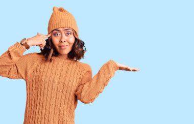 Yün kazak ve kış şapkası giyen genç, güzel, melez bir kadın. Avuç içinin açık olması, fotokopi alanını göstermesi ve alnını işaret etmesi kafasını karıştırıyor. Bunu bir düşün.. 