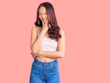 Gündelik kıyafetler giyen genç, güzel Çinli kız kameraya bakıyor kollarını kavuşturup gülümsüyor ve çenesini kaldırıyor. Olumlu düşünmek. 