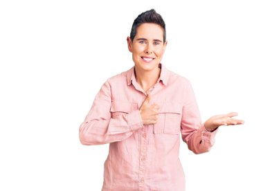 Kısa saçlı, günlük kıyafetler giyen, avuçlarını gösteren ve baş parmaklarıyla hareket eden mutlu ve neşeli gülümseyen genç bir kadın. 