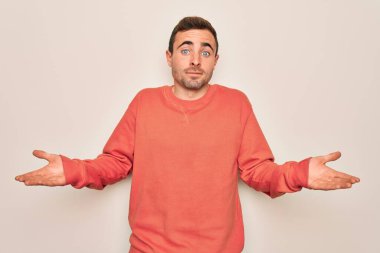 Mavi gözlü, beyaz arka planda dikilen sıradan süveter giyen, elleri ve kolları havada, kafası karışmış genç, yakışıklı bir adam. Şüphe kavramı.